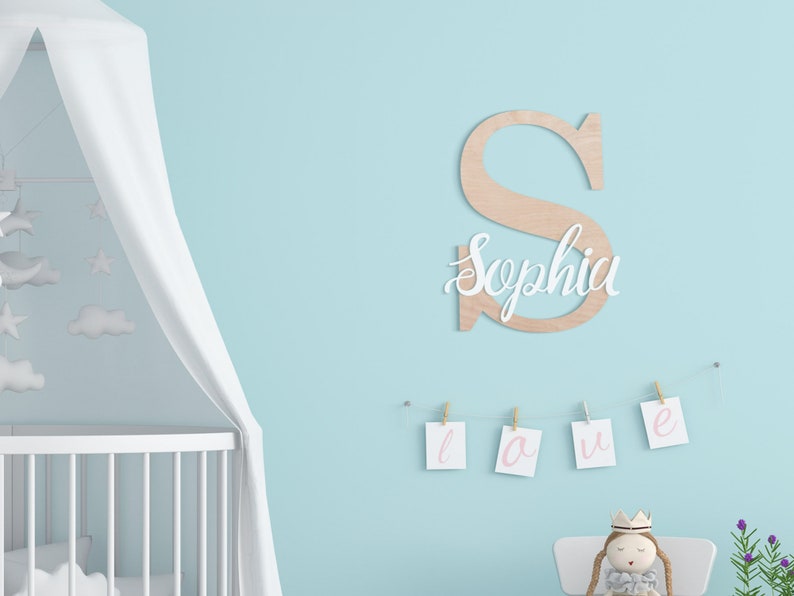 K&ouml;nnte beinhalten: Ein wei&szlig;er Holzbuchstabe S mit dem Namen Sophia in wei&szlig;er Kursivschrift auf der Vorderseite. Der Buchstabe h&auml;ngt an einer hellblauen Wand mit einem wei&szlig;en Kinderbett und einem wei&szlig;en Baldachin im Hintergrund. Unter dem Buchstaben h&auml;ngt eine Schnur mit wei&szlig;en Karten, auf denen das Wort "love" in rosa Kursivschrift steht.
