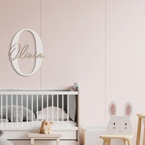 K&ouml;nnte beinhalten: Wei&szlig;er Holzbuchstabe O mit dem Namen Olivia in Kursivschrift darauf. Der Buchstabe ist an einer hellrosa Wand &uuml;ber einem wei&szlig;en Kinderbett mit einer grau-wei&szlig;en gemusterten Matratze angebracht. Ein wei&szlig;er Stuhl in Form eines Kaninchens befindet sich im Vordergrund.