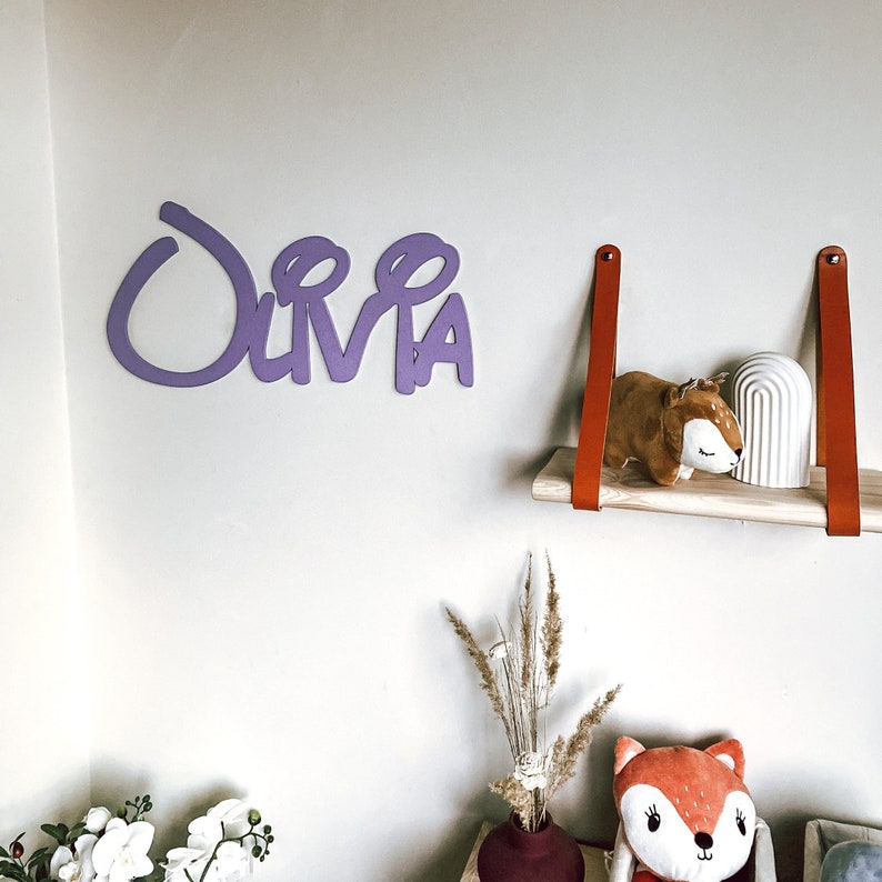May include: A purple wooden name sign spelling "Olivia" with a whimsical font. The sign is mounted on a white wall. A wooden shelf with two brown leather straps hangs below the sign. A small stuffed animal and a white ceramic object are on the shelf.