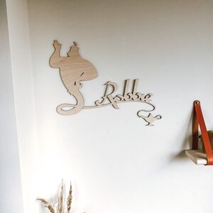 May include: A wooden wall decoration featuring a silhouette of the Genie from Aladdin, the name "Robbie" in a whimsical font, and a small lamp. The decoration is mounted on a white wall.
