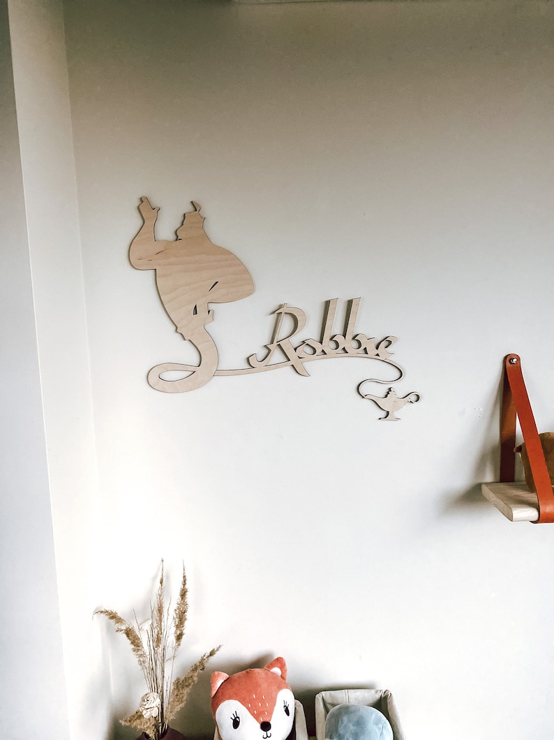 May include: A wooden wall decoration featuring a silhouette of the Genie from Aladdin, the name "Robbie" in a whimsical font, and a small lamp. The decoration is mounted on a white wall.