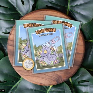 Könnte beinhalten: Drei Mockup-Bilder eines lila und blauen Drachen-Schlüsselanhängers mit floralem Design. Der Schlüsselanhänger ist mit "Taschen-Talisman" und "Artwork von Pewy" beschriftet.
