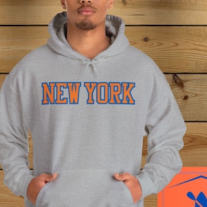 May include: A gray hooded sweatshirt with the text "NEW YORK" in orange and blue lettering. The sweatshirt is being modeled by a person with short curly hair.