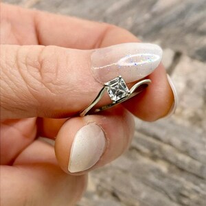 May include: A silver ring featuring a square-cut, clear gemstone. The ring's band has a curved design, and the gemstone is held in place by four prongs. The ring is set against a blurred wooden backdrop.