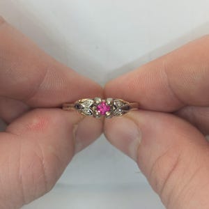 May include: A gold ring featuring a central square-cut vibrant pink gemstone held in a four-prong setting. The band is accented with small, clear stones embedded in decorative metalwork on both sides of the center stone, creating a detailed design.