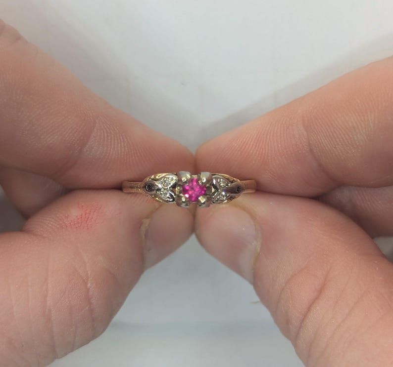 May include: A gold ring featuring a central square-cut vibrant pink gemstone held in a four-prong setting. The band is accented with small, clear stones embedded in decorative metalwork on both sides of the center stone, creating a detailed design.