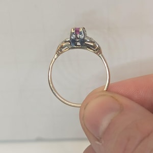 May include: A gold ring featuring a central prong-set pink gemstone. The band is thin and circular, with decorative metalwork extending from the sides of the setting toward the shoulders of the ring, showcasing a polished metallic finish.