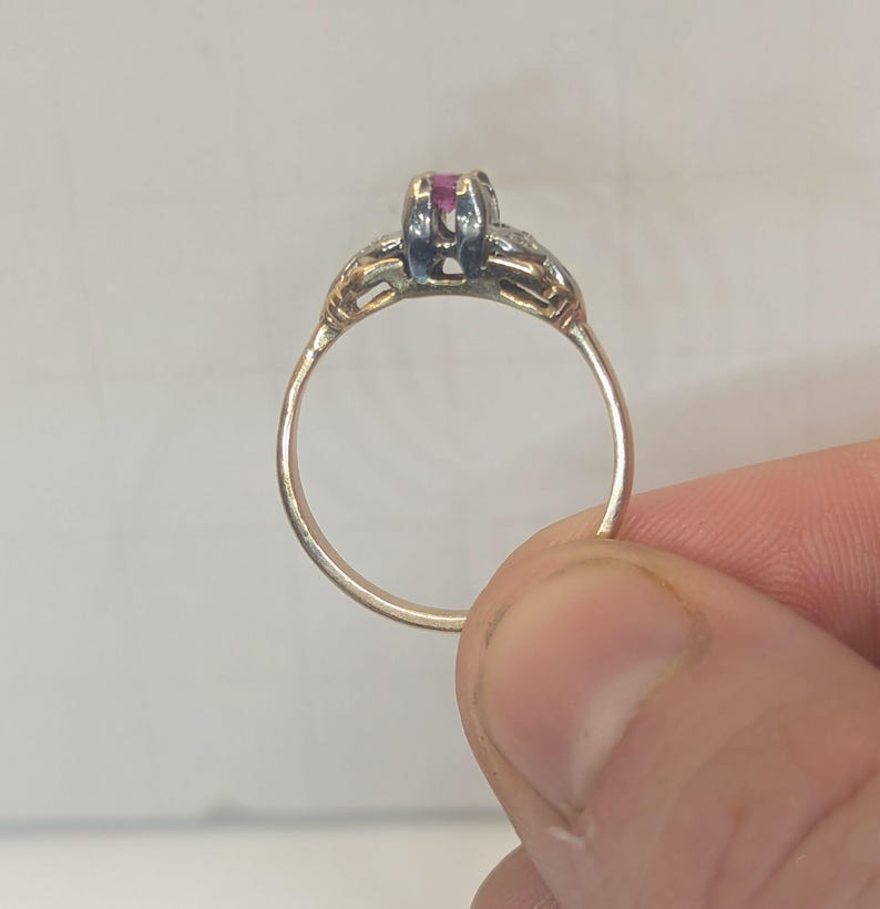 May include: A gold ring featuring a central prong-set pink gemstone. The band is thin and circular, with decorative metalwork extending from the sides of the setting toward the shoulders of the ring, showcasing a polished metallic finish.