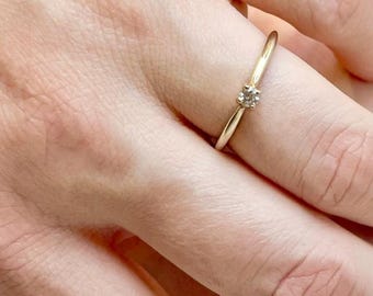 Op de afbeelding: Een delicate gouden ring met een kleine, heldere edelsteen in het midden. De ring heeft een dunne band en een eenvoudig, elegant ontwerp. De edelsteen is rond en lijkt te zijn gezet in een vier-klauwzetting. De ring zit om een vinger.