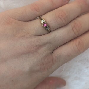 May include: A gold ring featuring a central round pink gemstone flanked by two small clear stones. The metal band has an intricate, engraved design on both sides of the setting, showcasing a delicate and detailed metalwork pattern throughout the band.