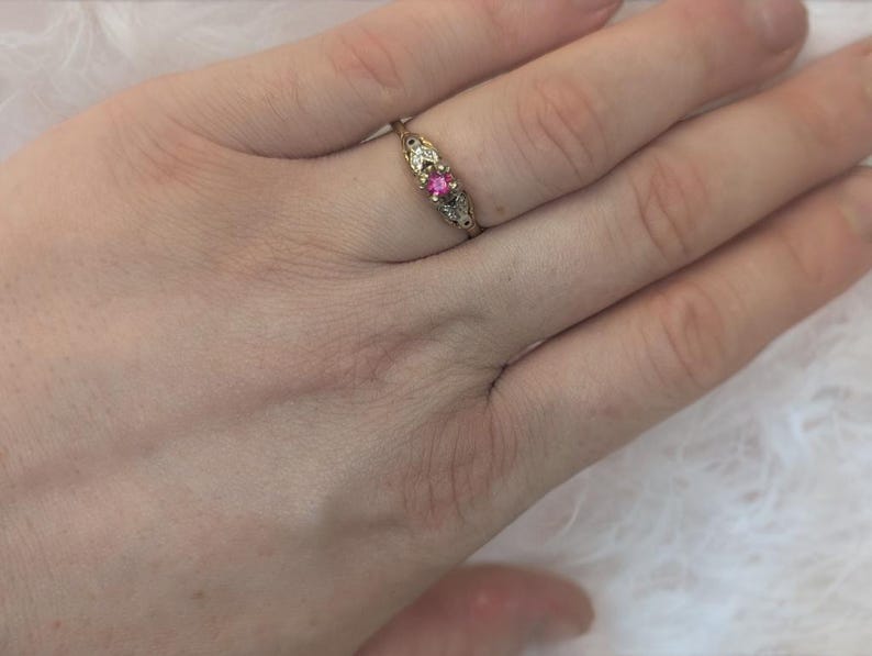 May include: A gold ring featuring a central round pink gemstone flanked by two small clear stones. The metal band has an intricate, engraved design on both sides of the setting, showcasing a delicate and detailed metalwork pattern throughout the band.