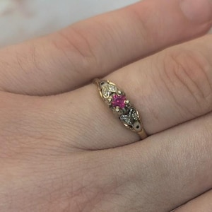 Könnte beinhalten: Ein goldener Ring mit einem zentralen, quadratisch geschliffenen, leuchtend pinkfarbenen Edelstein, der von zwei kleineren klaren Steinen flankiert wird, die in eine dekorative Metallfassung eingelassen sind, welche sich zum Ringband hin verjüngt.