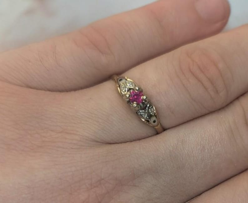 May include: A gold ring featuring a central square-cut vibrant pink gemstone flanked by two smaller clear stones set within a decorative metal mounting that tapers along the band, showcasing a detailed design on the shoulders of the ring.