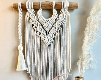 May include: A white macrame wall hanging with a wooden dowel at the top. The hanging features a geometric pattern with a tassel on the left side.