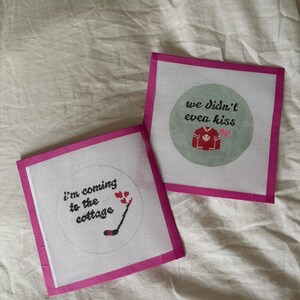 May include: Two needlepoint canvases with pink borders. One canvas reads "I'm coming to the cottage" with a hockey stick and hearts. The other says "we didn't even kiss" with a hockey jersey and hearts.