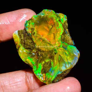May include: A raw, unpolished opal gemstone displaying vibrant flashes of neon green, orange, and yellow play-of-color against a dark, earthy matrix. The stone features an irregular, textured surface with the text CabochonCollection21 visible.