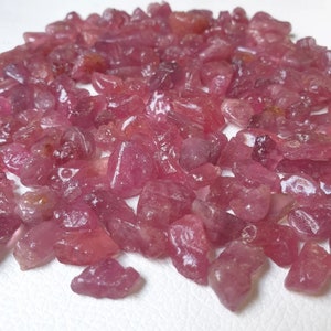 May include: A pile of pink ruby gemstone chips. The chips are various sizes and shapes, with some having a more pointed shape and others being more rounded.