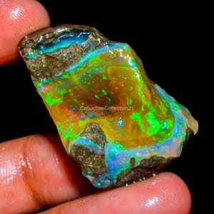 May include: A raw, unpolished opal gemstone featuring a vibrant play of color with flashes of neon green, orange, and blue against a translucent amber base. The stone has rough, textured grey matrix inclusions and the text CabochonCollection21.