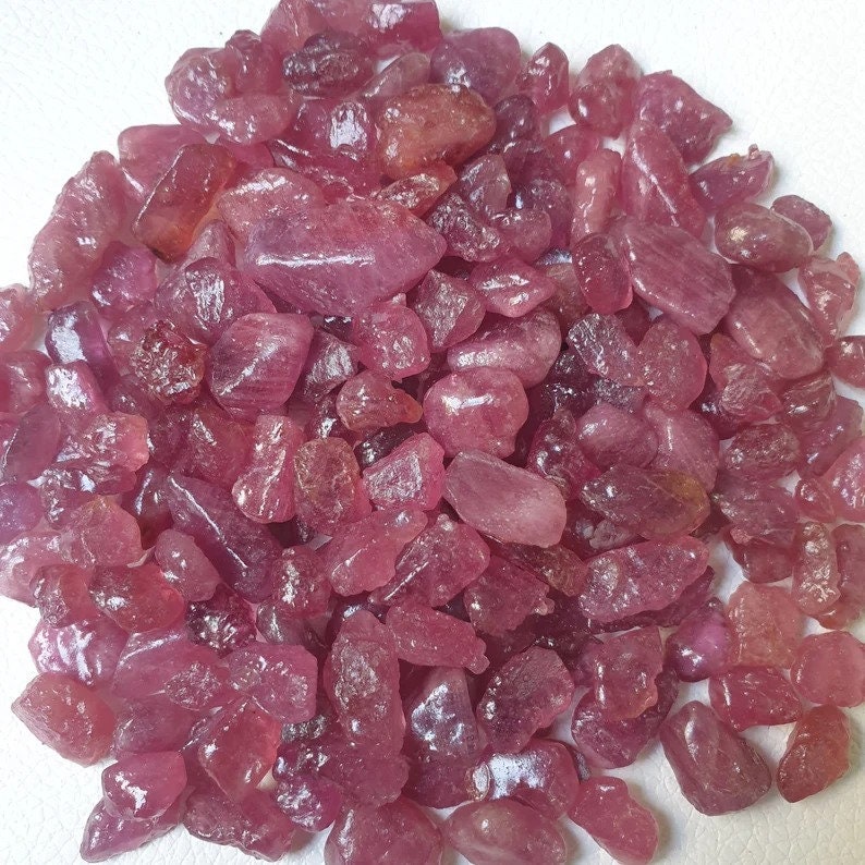 May include: A pile of loose pink ruby gemstones. The rubies are various sizes and shapes, with some having a more pointed shape and others being more rounded.