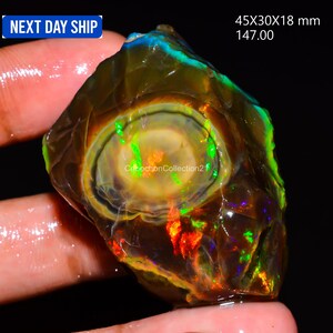 May include: A raw, dark brown opal gemstone featuring a central circular pattern with vibrant flashes of neon green, orange, and blue play-of-color. The stone measures 45x30x18 mm and displays a polished, translucent surface with natural inclusions.