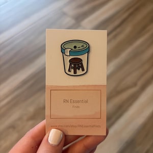 May include: A white card with a cartoon pin of a stool inside a container. The pin has a smiling face. Below the pin is a brown rectangle with the text "RN Essential Finds" and a website address.