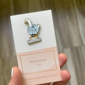 May include: A metal pin featuring a cartoon goose wearing a light blue medical coat and stethoscope. The pin has the text "TRUST ME I'M A DUCKCTOR" on a white background. The pin is on a white card with a pink label that says "RN Essential Finds".