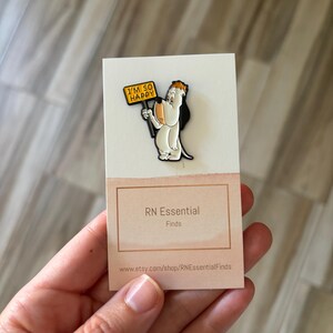 May include: A colorful enamel pin featuring a cartoon dog holding a sign that reads "I'M SO HAPPY". The pin is attached to a white card with the text "RN Essential Finds".