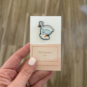 May include: A novelty pin featuring a cartoon goose wearing a nurse's cap. The goose is light blue with white accents and the text "registered silly goose". The pin is on a white card with the text "RN Essential Finds".