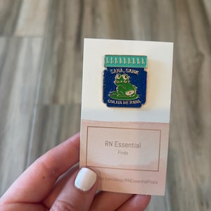 May include: A blue and green enamel pin featuring a frog inside a jar with the words "Sana Sana" and "Colita de Rana". The pin is attached to a white card with the text "RN Essential Finds".