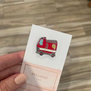 May include: A red and silver fire truck enamel pin with the number 1 and a yellow 9 on the side. The pin is on a white card with the text "RN Essential Finds".