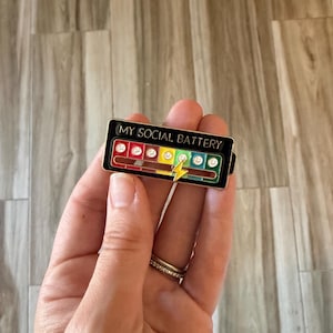 May include: A rectangular enamel pin with the text "MY SOCIAL BATTERY" at the top. The pin features a battery level indicator with colorful segments and a lightning bolt graphic. The battery level is low.