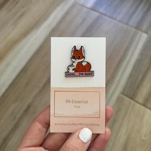 May include: A pin featuring a cartoon fox with orange and white fur, holding a mug. The pin has the text "SHHH... I'M BUSY" on a pink banner. The pin is on a white and tan card with the text "RN Essential Finds".