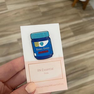 May include: Blue and white enamel pin of a medicine bottle with the text "Sana Sana" and "Colonia de Rom" on it. The pin is on a white card with the text "RN Essential Finds" and a website address.