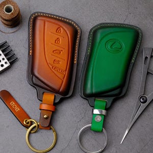 May include: Two leather key fobs for Lexus vehicles, one in a gradient brown and one in a solid green, each featuring embossed button icons and a logo. The brown fob includes a small leather tag with the name BOB and a brass-toned metal ring.