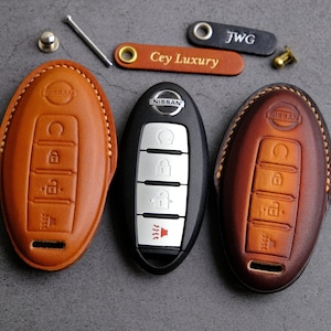Puede incluir: Tres fundas de cuero para llaves de coche Nissan. Las fundas de llave son marrones, beige y marrón oscuro. Las fundas de llave tienen el logotipo de Nissan y botones para bloquear, desbloquear y abrir el maletero.