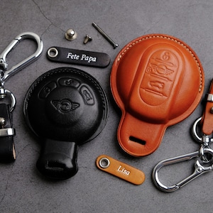 Può includere: Due portachiavi in ​​pelle con targhette personalizzate. Un portachiavi è nero con un logo Mini Cooper e l'altro è marrone con un logo Mini Cooper. I portachiavi sono su una superficie grigia.