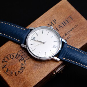 May include: A silver wristwatch with a white face and Roman numeral hour markers. The watch has a blue leather band with a light brown stitching detail. The watch is sitting on a wooden box with the text "MP HABET se & symbols (pcs)" engraved on the top.