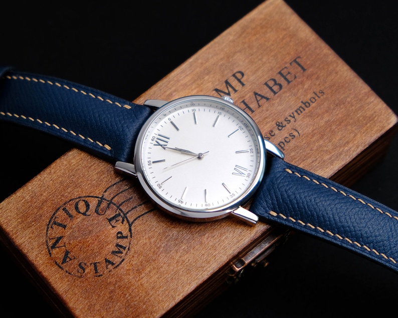 May include: A silver wristwatch with a white face and Roman numeral hour markers. The watch has a blue leather band with a light brown stitching detail. The watch is sitting on a wooden box with the text "MP HABET se & symbols (pcs)" engraved on the top.