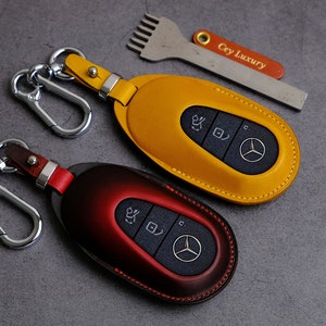 May include: Two leather key fobs with metal key rings and car key covers. The key fobs are yellow and red. The car key covers have a Mercedes-Benz logo and buttons. The text "Cey Luxury" is visible on a metal tool.