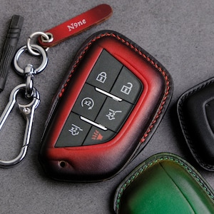 May include: A red leather key fob with stitching around the edges. The fob has five buttons with icons for lock, unlock, trunk release, and panic. The fob is attached to a silver key ring with a small screwdriver and a leather strap with the word "None" engraved on it.