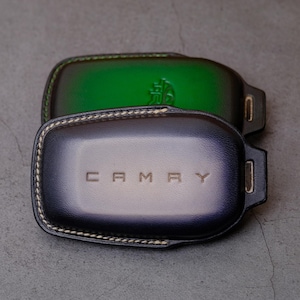 May include: Two leather key fob covers, one green and one dark blue. The dark blue cover has the word "CAMRY" embossed on it. Both covers have a stitched border and a small metal loop.