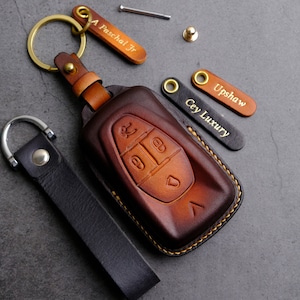 May include: A handcrafted leather key fob and key holder set in shades of brown and black. The key fob features embossed buttons and a gold-colored keyring. Includes personalized leather tags with text.