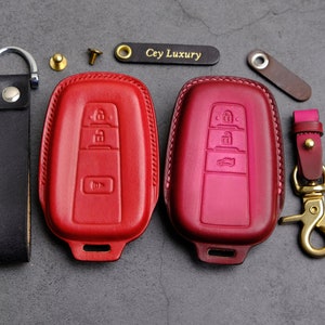 May include: Two red leather key covers with stitching detail. The key covers have three buttons each and are designed for car keys. The text "Cey Luxury" is printed on a small gold tag.