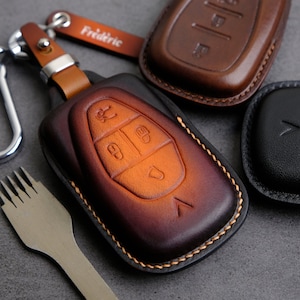May include: Three leather key covers, one brown, one orange, and one black. The brown cover has the word "Freidric" embossed on it. The orange cover has a design of four buttons and a chevron. The black cover has a chevron design.