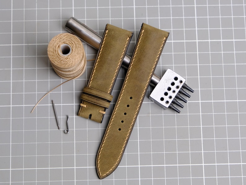 May include: Olive green leather watch straps with cream stitching, alongside a spool of thread, needles, and tools. The straps have a classic design with multiple holes for adjustment. The tools suggest a handmade or repair theme.