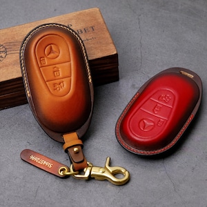 May include: Two leather car key cases in shades of brown and red. The brown case has a key ring and a gold-colored clasp. Both cases feature embossed key symbols and a car logo.