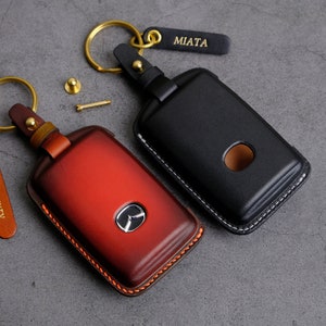 May include: Two leather keychains with metal rings. One keychain is black with the word "MIATA" on a tag. The other keychain is red with a logo on the front. Both keychains have stitching around the edges.