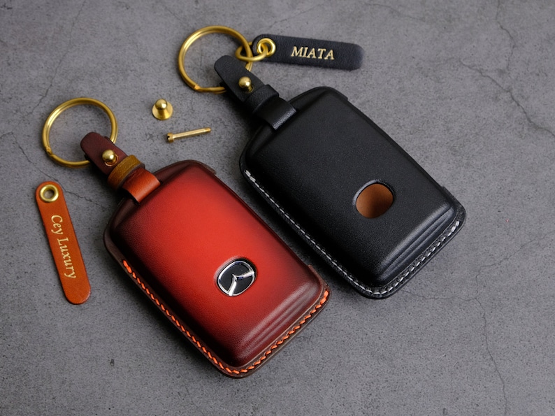 May include: Two leather keychains with metal rings. One keychain is black with the word "MIATA" on a tag. The other keychain is red with a logo on the front. Both keychains have stitching around the edges.