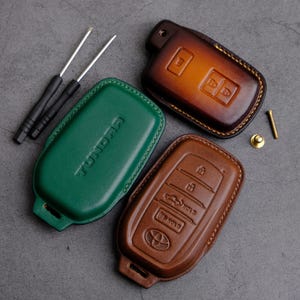 Puede incluir: Tres fundas de cuero para llaves de coche. Una es verde y dice "TUNDRA". Otra es marrón con un diseño de cuatro botones. La última es marrón con un diseño de dos botones.