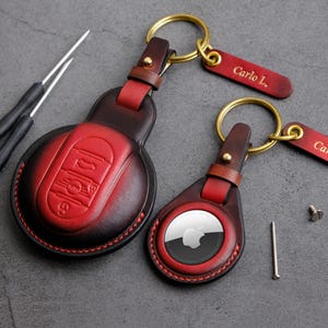 Può includere: Due portachiavi in ​​pelle con accenti rossi e neri. I portachiavi presentano un portachiavi rotondo in pelle cucito con un logo e il testo "Carlo L." su un'etichetta in pelle separata.